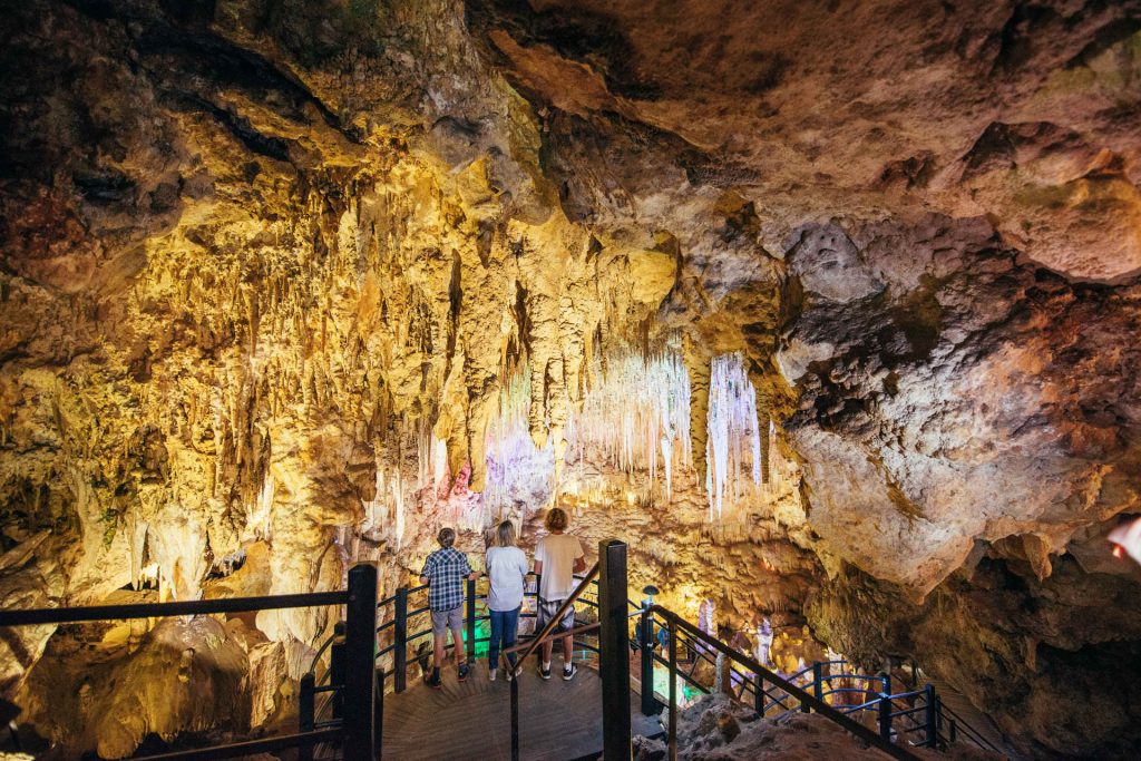 3 people standing on a wooden landing and looking at the cave formations. A coloured light is shining on the stalagmites hanging from the cave ceiling.