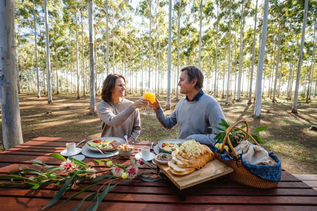A man and woman about to eat breakfast on the front deck of the chalet. They are holding flutes of orange juice and toasting each other. The view of the trees can be seen in the distance.