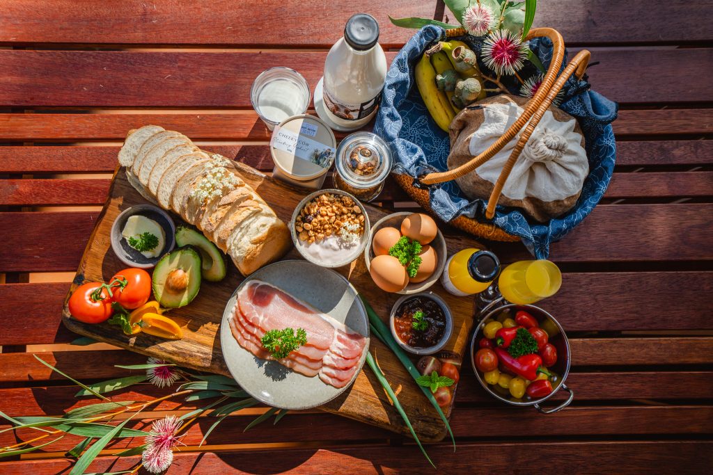 An overhead view of the complimentary breakfast hamper. You can see bacon on a plate, eggs, orange juice, bread on a bread board, yoghurt and muesli in a bowl, chutney, tomatoes, avocado, butter and milk.