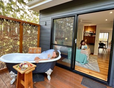 Image 11: The back deck of the chalet with a girl having a bubble bath. In the bedroom there is another girl laying on the bed reading a book.