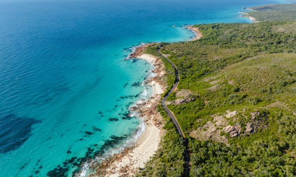 An overhead drone shot of a road following the coast. The coastline is rocky but the sea is calm. The vegetation is low lying and very green.