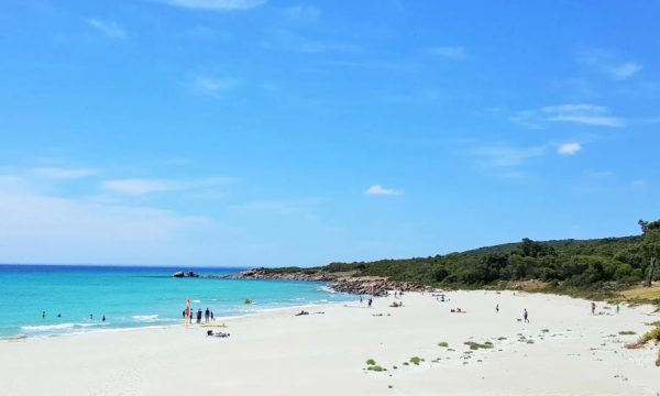 A local beach in Busselton. There is a large section of white sand with people sitting and laying on. The water is calm. In the background there is a rocky section of the beach