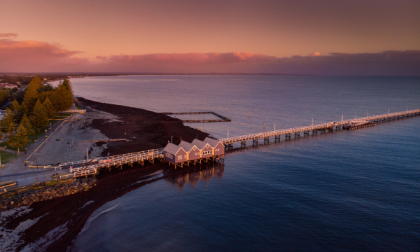 A wooden jetty in Busselton at sunset. The sky is pink. There is another smaller U-shaped jetty in the background. The ocean is calm and blue. There is a lot of seaweed on the beach as it is winter. A building with 4 apex styled roofs is located at the beginning of the jetty. This is the visitor centre, where people can buy tickets to walk out to the end of the jetty or get a train to the end of the jetty which extends 1.8km out to sea